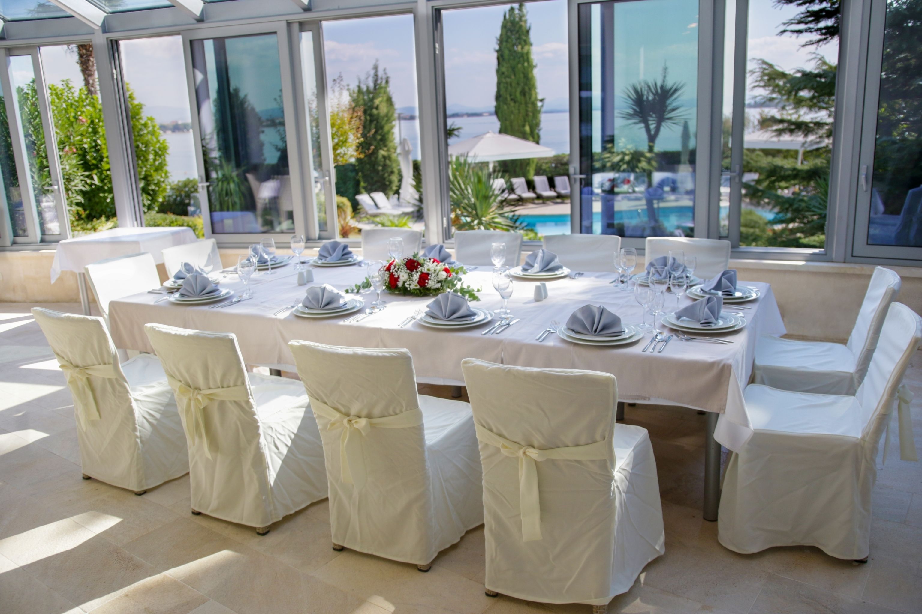 A dining area with a table set for six, surrounded by white chairs with bows, large windows, and a view of a pool and greenery.