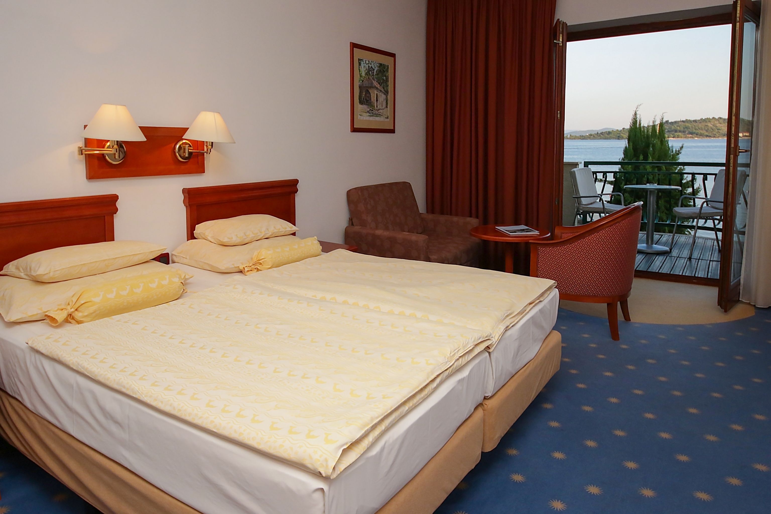 Une chambre d’hôtel avec un grand lit, deux lampes de chevet, une chaise, une petite table et un balcon avec vue sur la mer.
