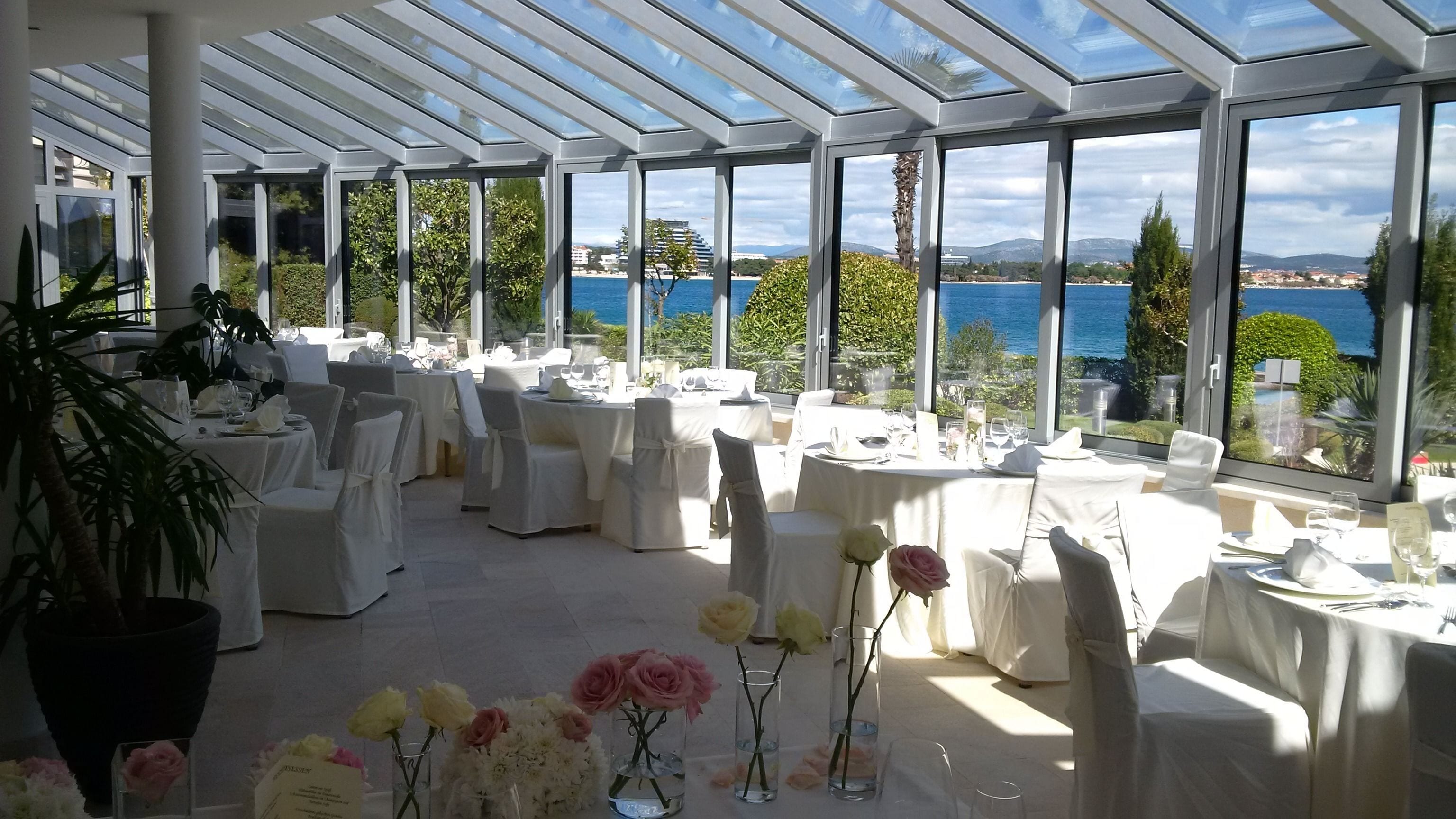 A sunlit outdoor dining area with tables set for a meal, overlooking a body of water and greenery.