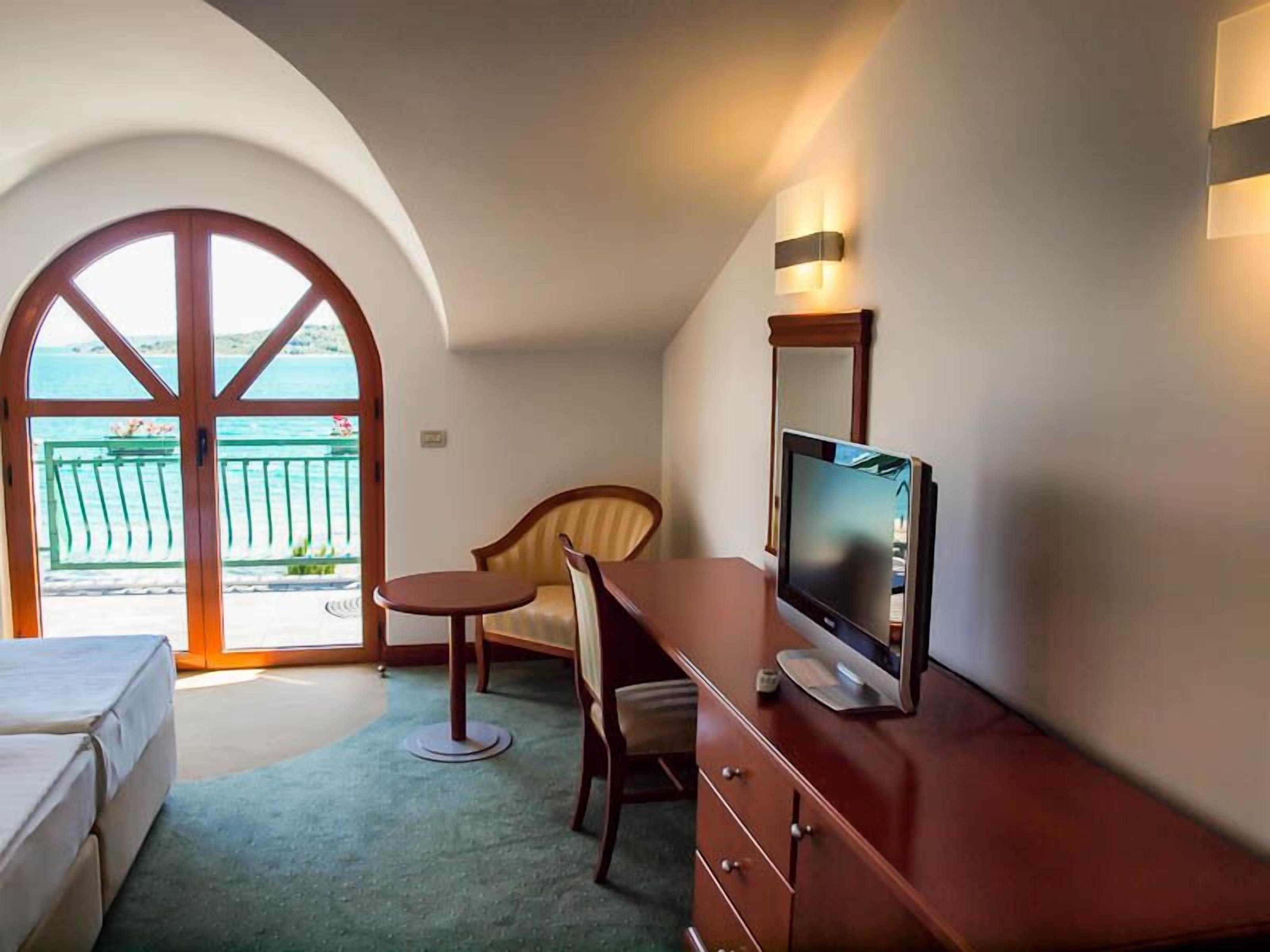 Ein Hotelzimmer mit großem Bogenfenster, einem Schreibtisch mit Fernseher, einem Sessel und einem Bett.