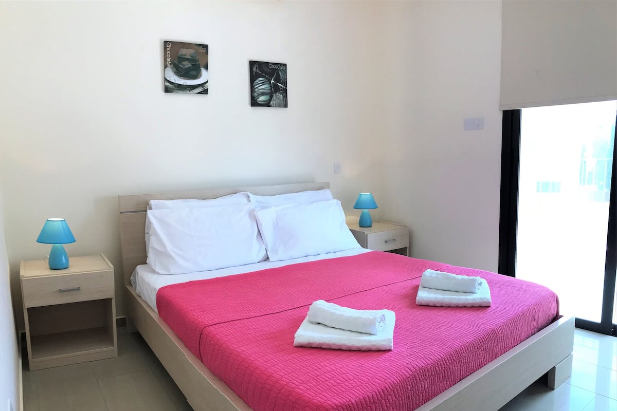 A neatly made bed with white pillows and a pink bedspread, two bedside tables with lamps, and two framed pictures on the wall.