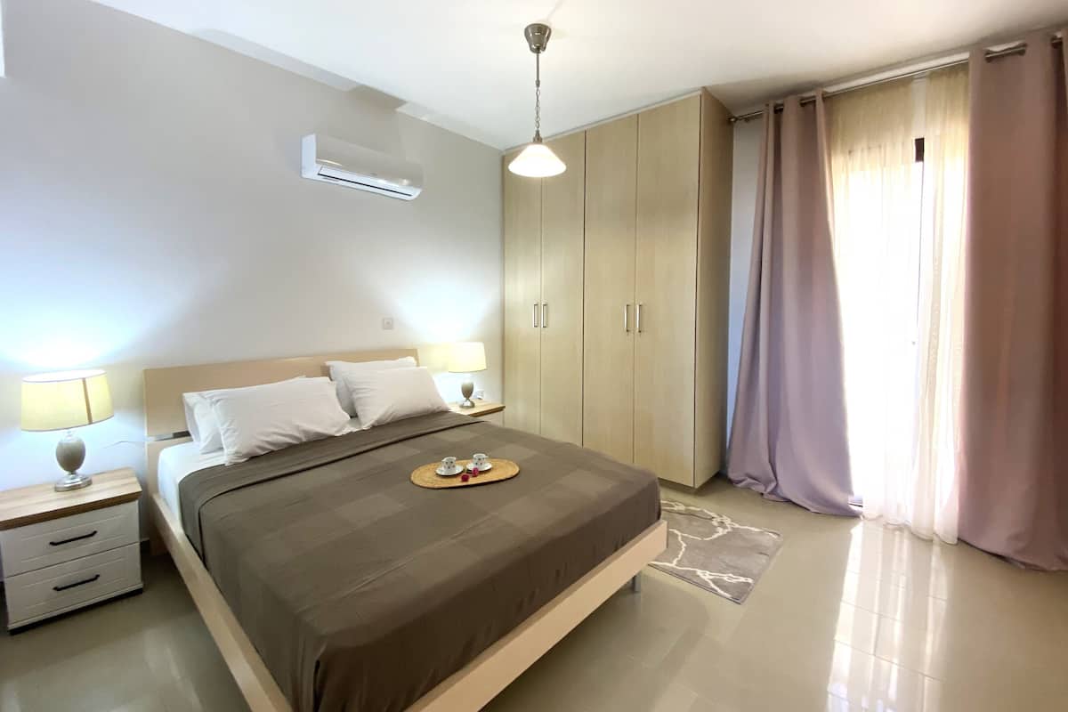 A bedroom with a bed, bedside tables, an air conditioning unit, and a window with curtains.