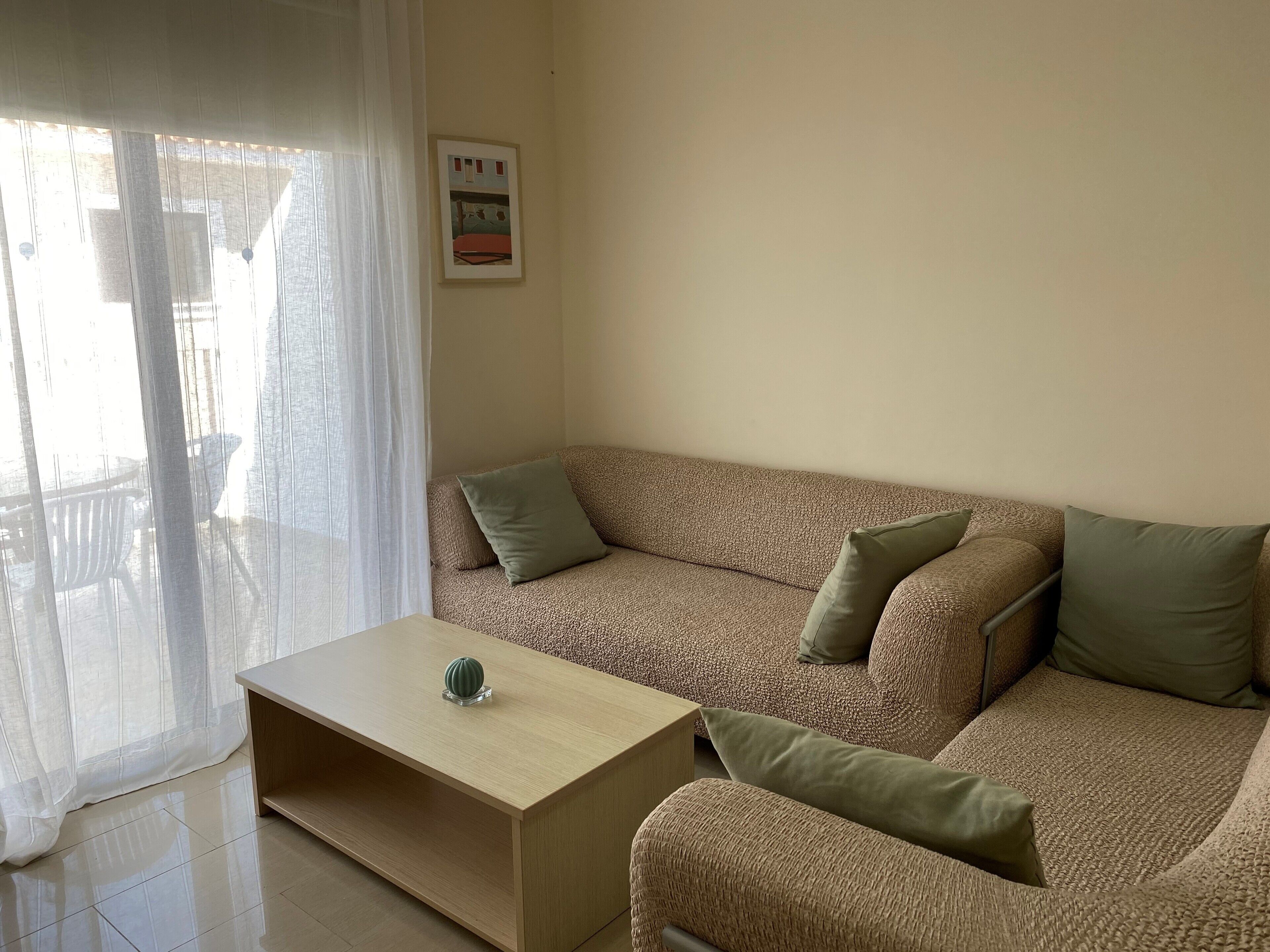 Un salón con un sofá color beige, dos cojines verdes, una mesa de centro de madera y un cuadro en la pared.