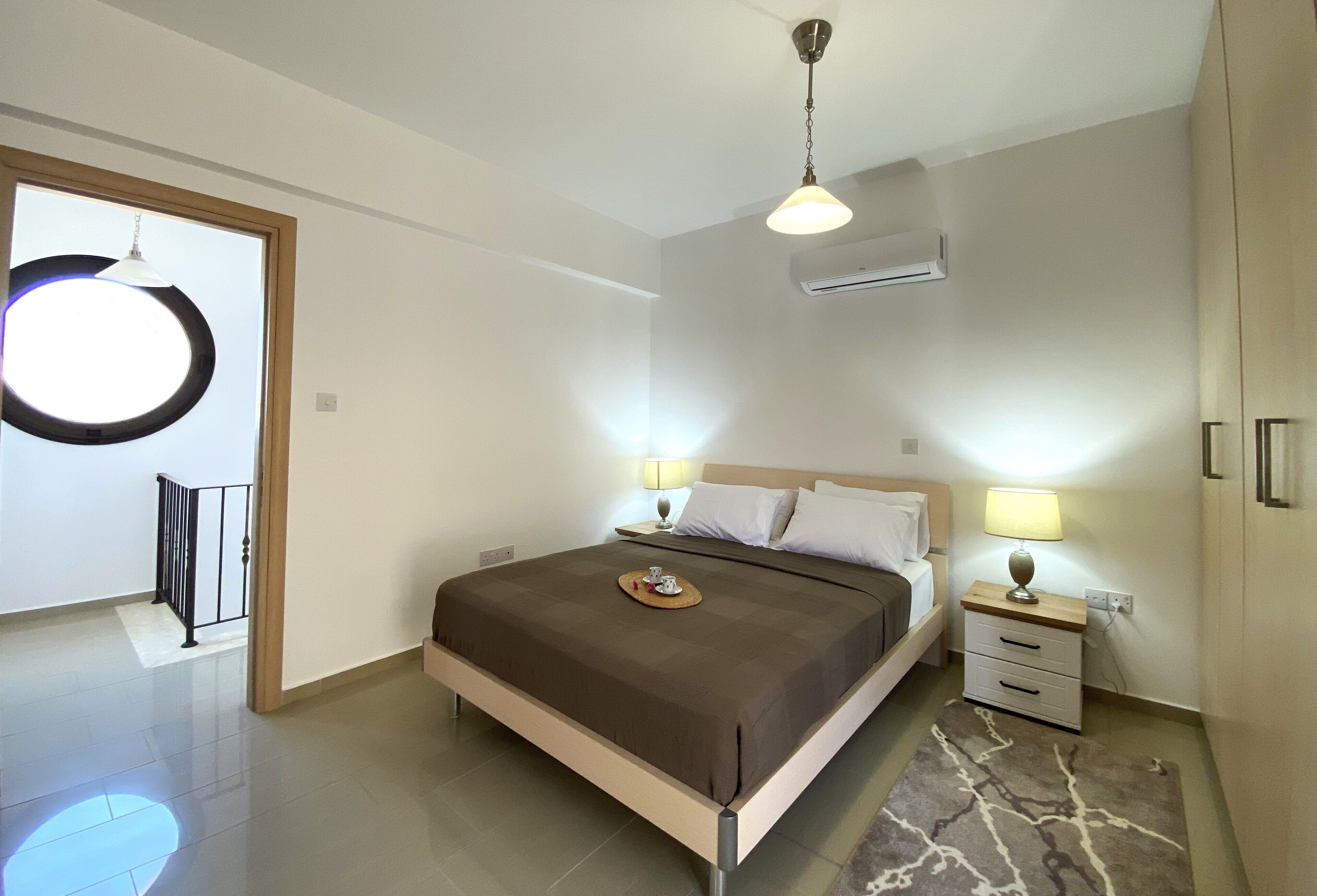A modern bedroom with a bed, bedside tables, a hanging light fixture, and an air conditioning unit.
