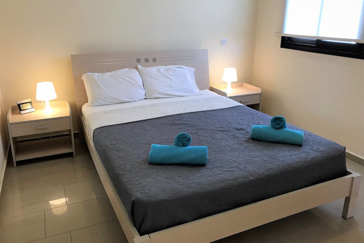 A neatly made bed with a grey bedspread and two blue towels, two bedside lamps, a white headboard, and a window with blinds.