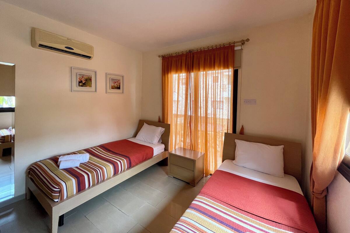 A room with two single beds, a small bedside table, a wall-mounted air conditioner, and a window with curtains.