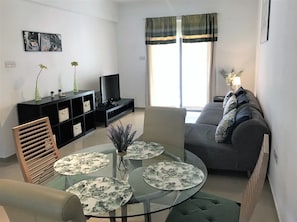 A living room with a glass table, sofa, TV, and a window with curtains. - Kyklades Resort & Spa (Paralimni)