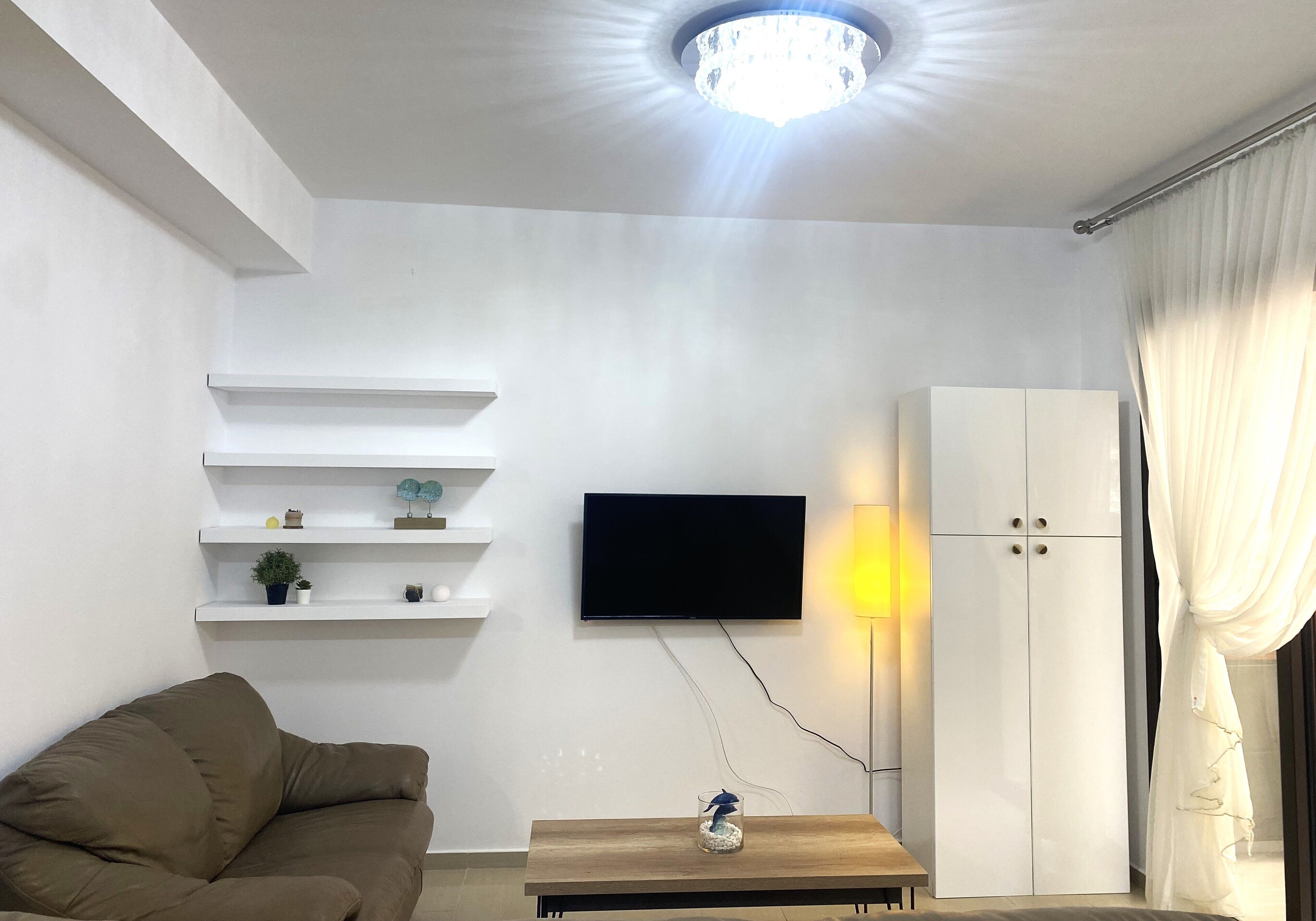 A modern living room with a flat-screen TV, white shelving, a brown sofa, a wooden coffee table, and a wardrobe.