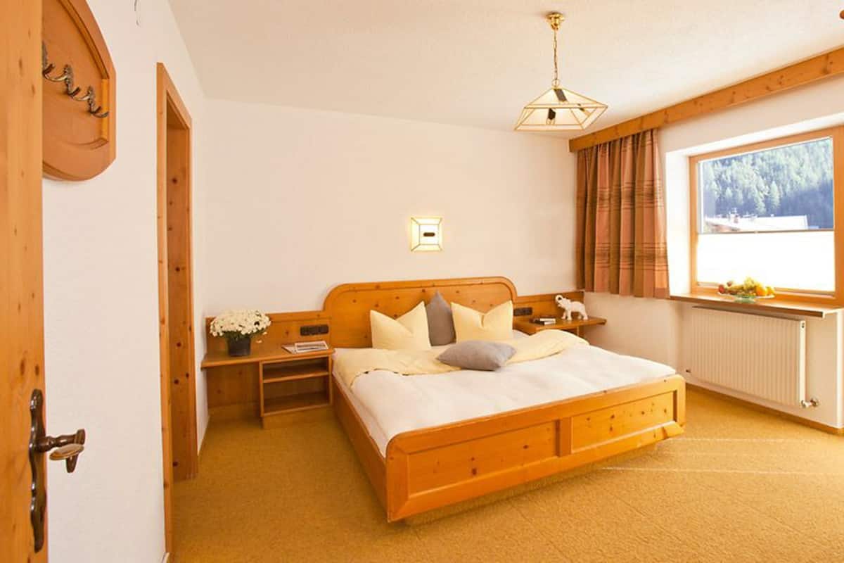 A bedroom with a wooden bed, a nightstand, a small table with a lamp, a window with curtains, and a radiator.