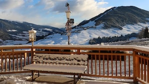 Un banco con asiento y respaldo cubiertos de nieve, baranda de madera y vista a un paisaje montañoso nevado con cielo despejado.
