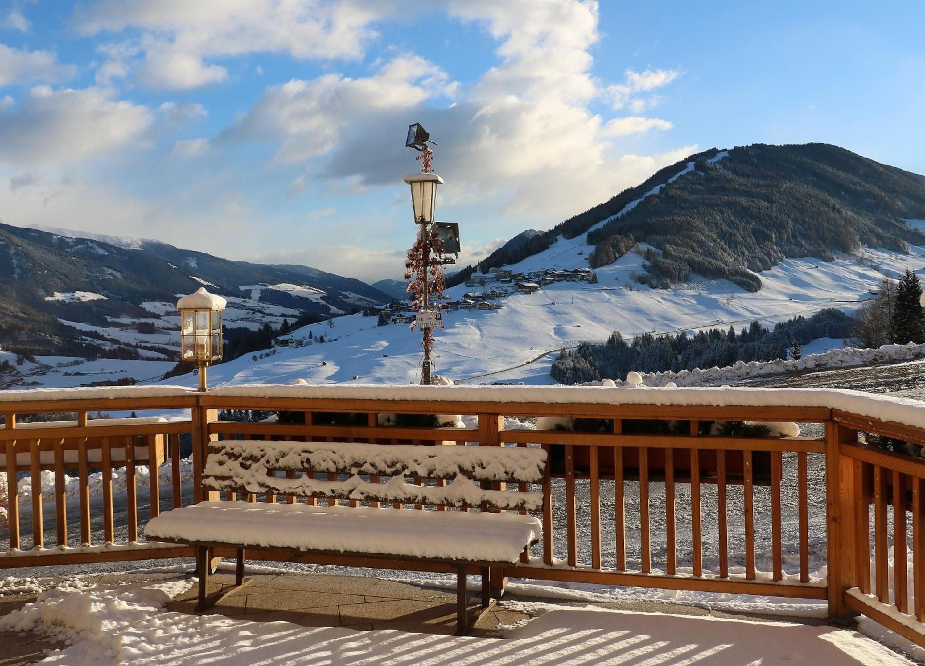 Un banc dont l’assise et le dossier sont recouverts de neige, avec une rambarde en bois, et qui offre une vue sur un paysage montagneux enneigé sous un ciel dégagé.