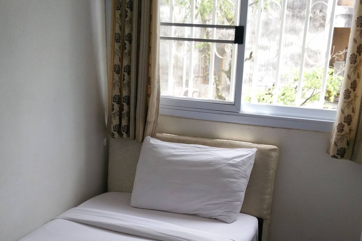 A small, neatly made bed with white linens in a room with a window, curtains, and a tiled floor.