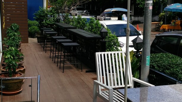 Un espace extérieur aménagé en terrasse, avec des tables et des chaises sous un grand parasol blanc, entouré de verdure et de voitures garées.