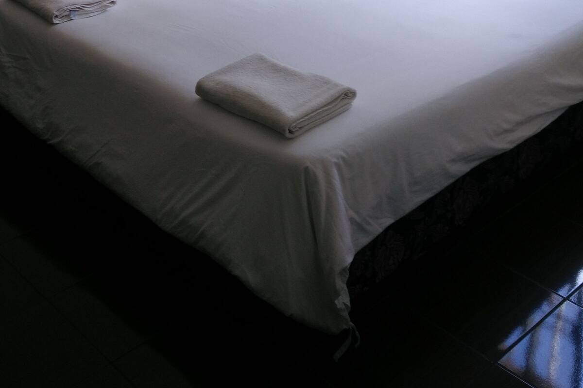 A neatly made bed with a folded towel, a wall clock, and a window with curtains.