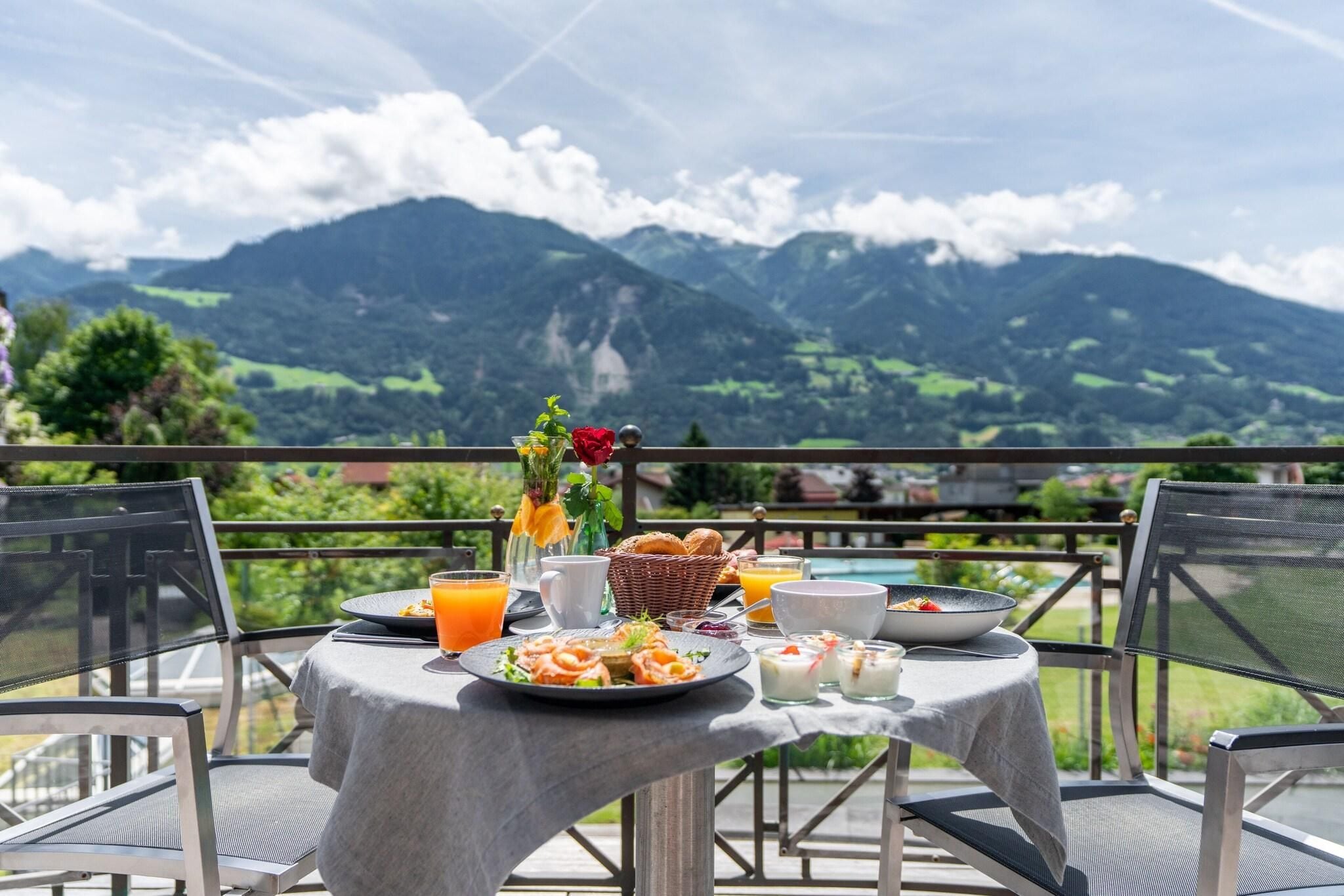 A table set for breakfast with a mountain view, including a basket of bread, a bowl of fruit, and a vase with flowers.