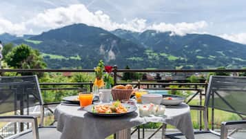 A table set for breakfast with a mountain view, including a basket of bread, a bowl of fruit, and a vase with flowers.