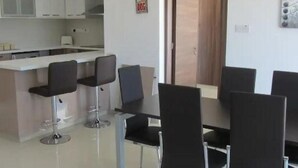A modern kitchen with white cabinets, a central island with bar stools, and a dining area with black chairs. - Bayview Gardens (Limassol)