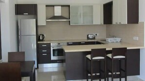 A modern kitchen with dark cabinets, a stainless steel refrigerator, a built-in oven, and a large island with bar stools. - Bayview Gardens (Limassol)