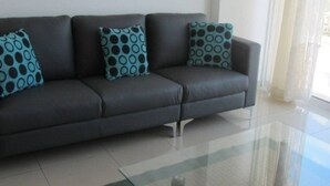 A dark grey sofa with three patterned cushions, a glass coffee table, and a white tiled floor. - Bayview Gardens (Limassol)