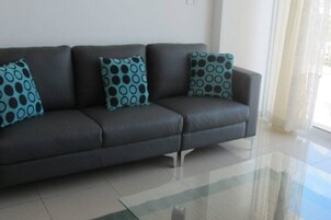 A dark grey sofa with three patterned cushions, a glass coffee table, and a white tiled floor.
