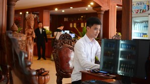A man seated at a wooden desk with a computer, in a room with ornate wooden furniture and a refrigerator stocked with beverages. - Holiday Hotel (Battambang)