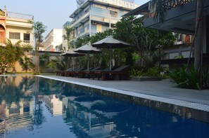 A swimming pool with lounge chairs and umbrellas, surrounded by greenery and buildings. - Holiday Hotel (Battambang)