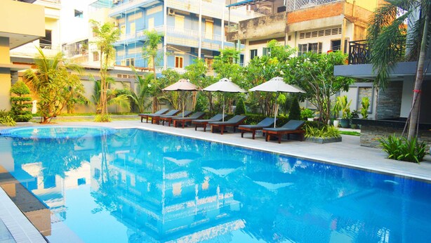 A swimming pool with lounge chairs and umbrellas, surrounded by greenery and buildings. - Holiday Hotel (Battambang)