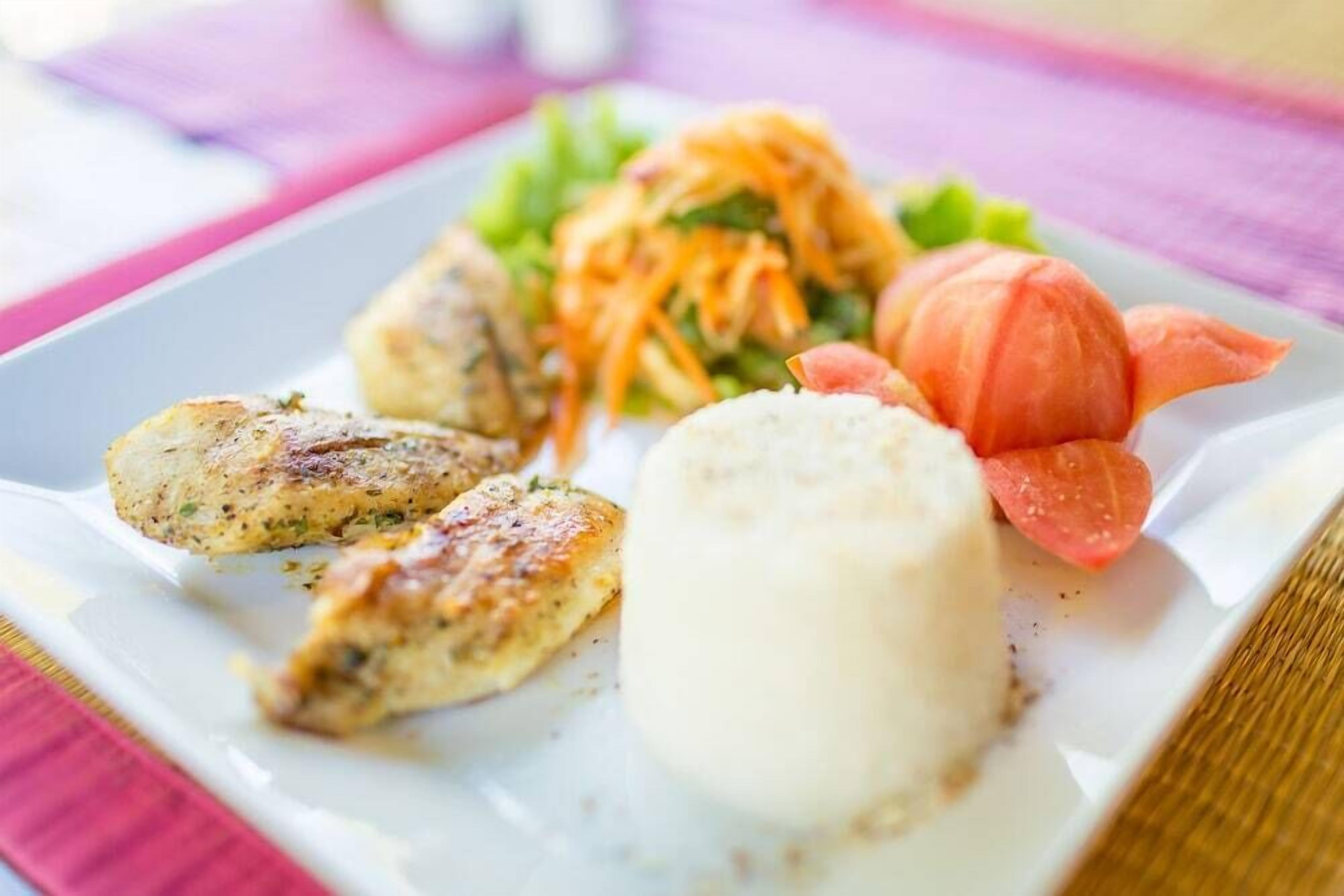 Ein Teller mit gegrilltem Hähnchen, einer Reisportion, einem Karotten Salat und einer Ziertomate.