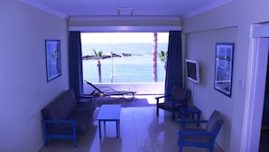 Quarto com sofá, cadeiras, mesa pequena, televisão e uma janela grande com vista para o mar e palmeiras.