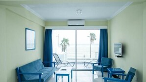 Quarto de hotel com sofá, duas cadeiras, uma mesa pequena e televisão. Há uma janela grande com vista para palmeiras e o mar.