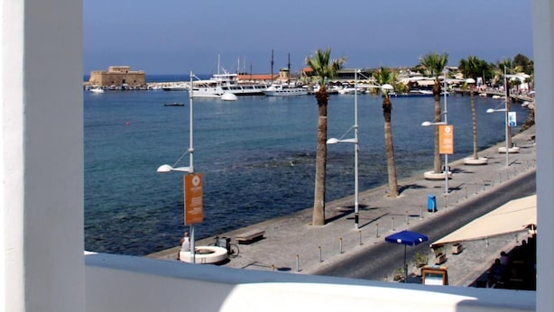 Vista costeira através de uma janela em arco, mostrando uma marina com barcos, uma praia arenosa com palmeiras e um edifício histórico ao fundo.