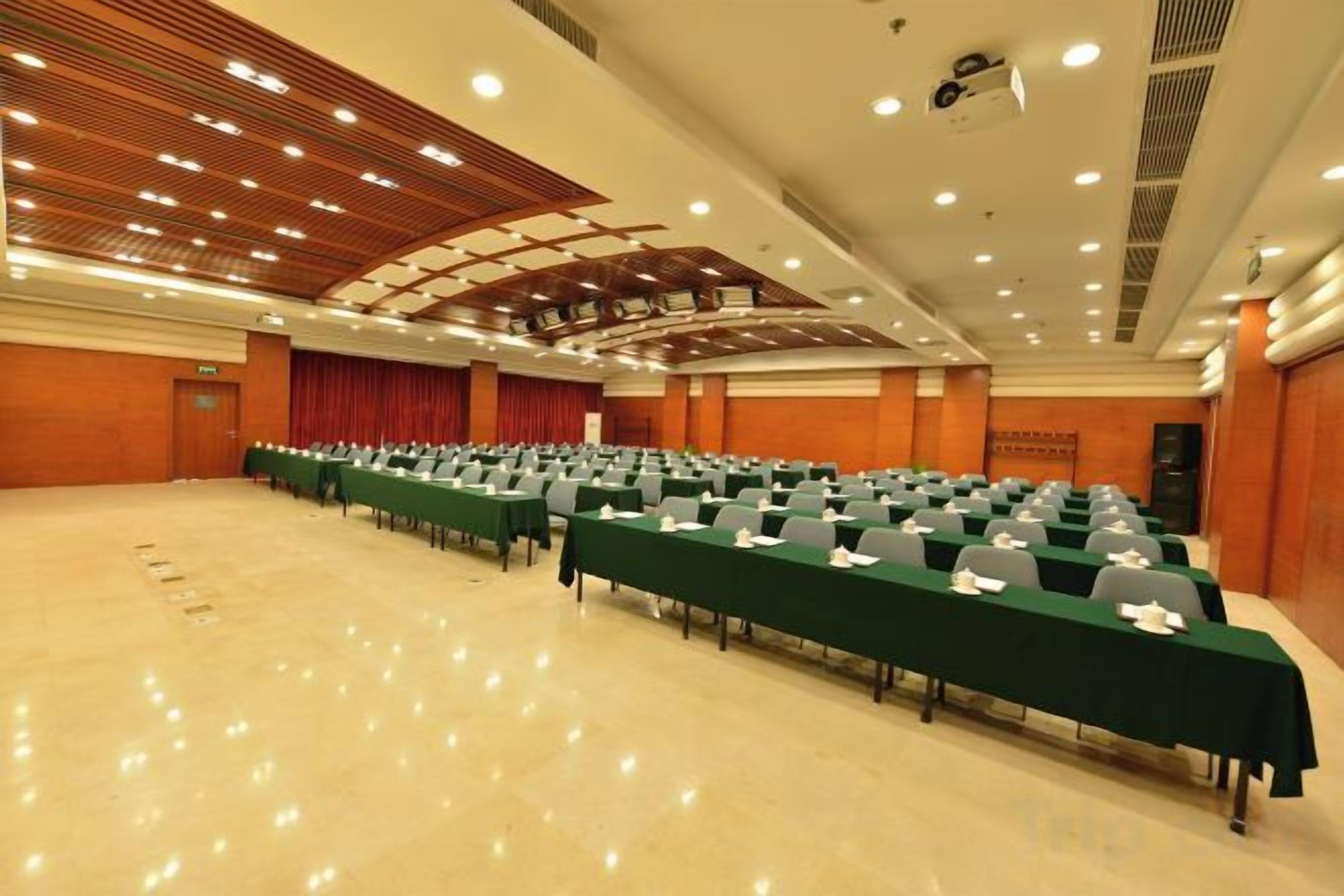 Un salón de conferencias con filas de mesas y sillas, un escenario con cortinas e iluminación empotrada en el techo.