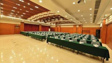 Un salón de conferencias con filas de mesas y sillas, un escenario con cortinas e iluminación empotrada en el techo.