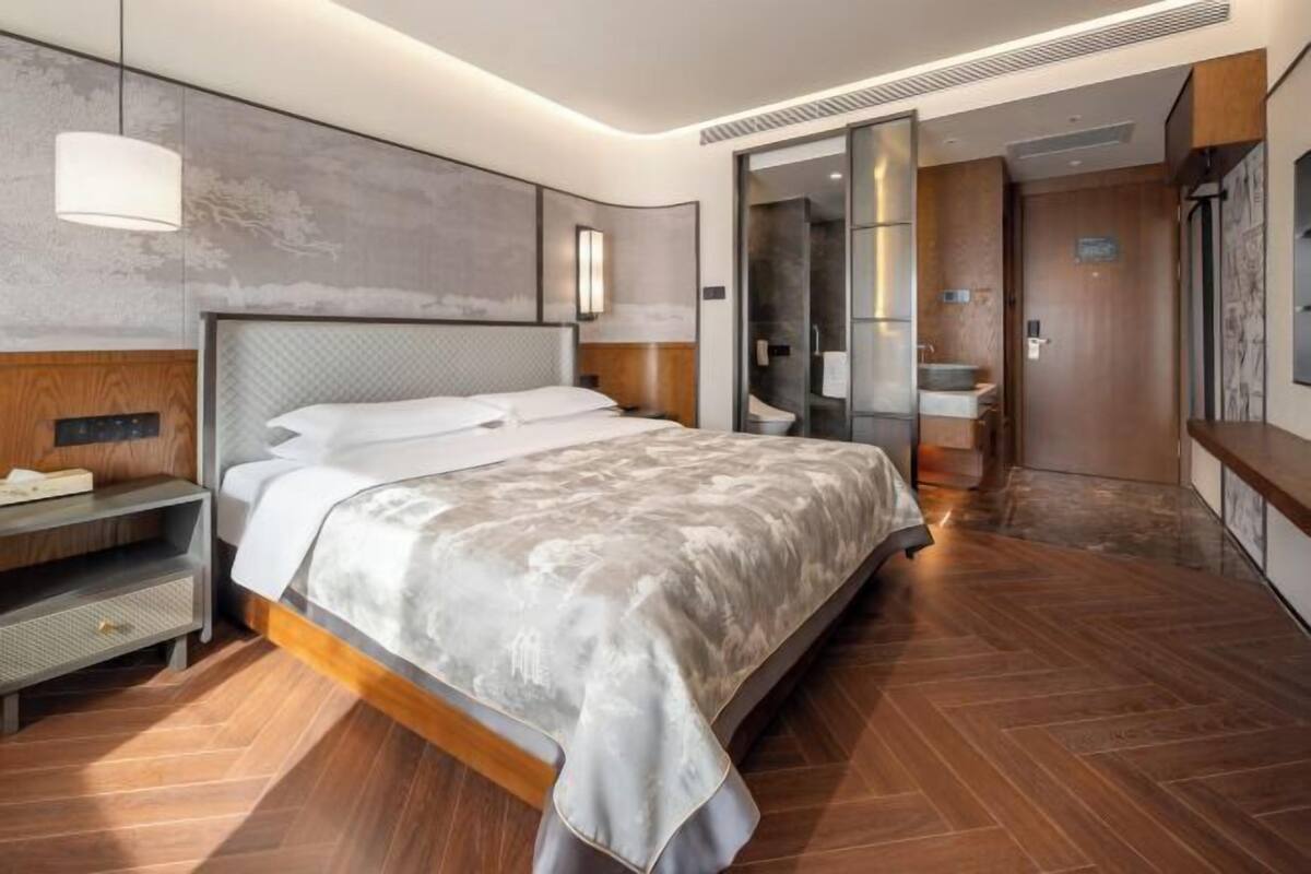 Une chambre d’hôtel moderne dotée d’un grand lit, d’une table de chevet, d’une armoire et d’une salle de bain visible à travers une porte en verre.