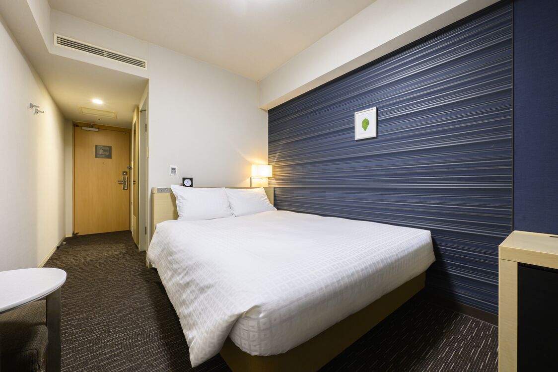 A hotel room with a bed, bedside table, lamp, and a door leading to another room.