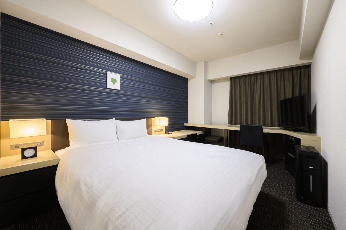 A hotel room with a bed, bedside lamps, a desk with a chair, a television, and a suitcase.