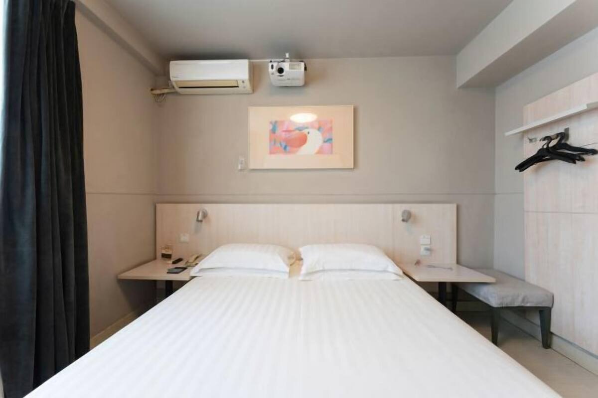 A hotel room with a bed, bedside tables, a desk, a chair, an air conditioner, a projector, and a wall-mounted coat rack.