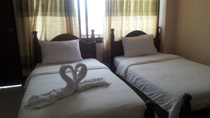 Two single beds with white linens and pillows, a heart-shaped towel arrangement on the left bed, and a window with curtains.