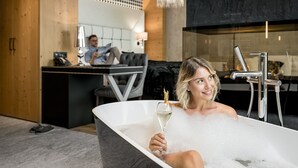 A person relaxing in a bathtub with a glass of wine, a modern bathroom with a large mirror, and a cozy seating area in the background.
