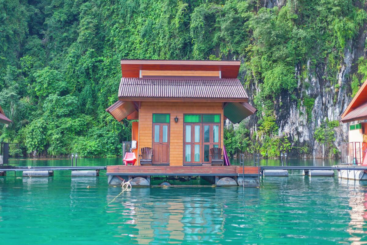 A wooden houseboat with a balcony, situated on calm water with a forested cliff in the background.