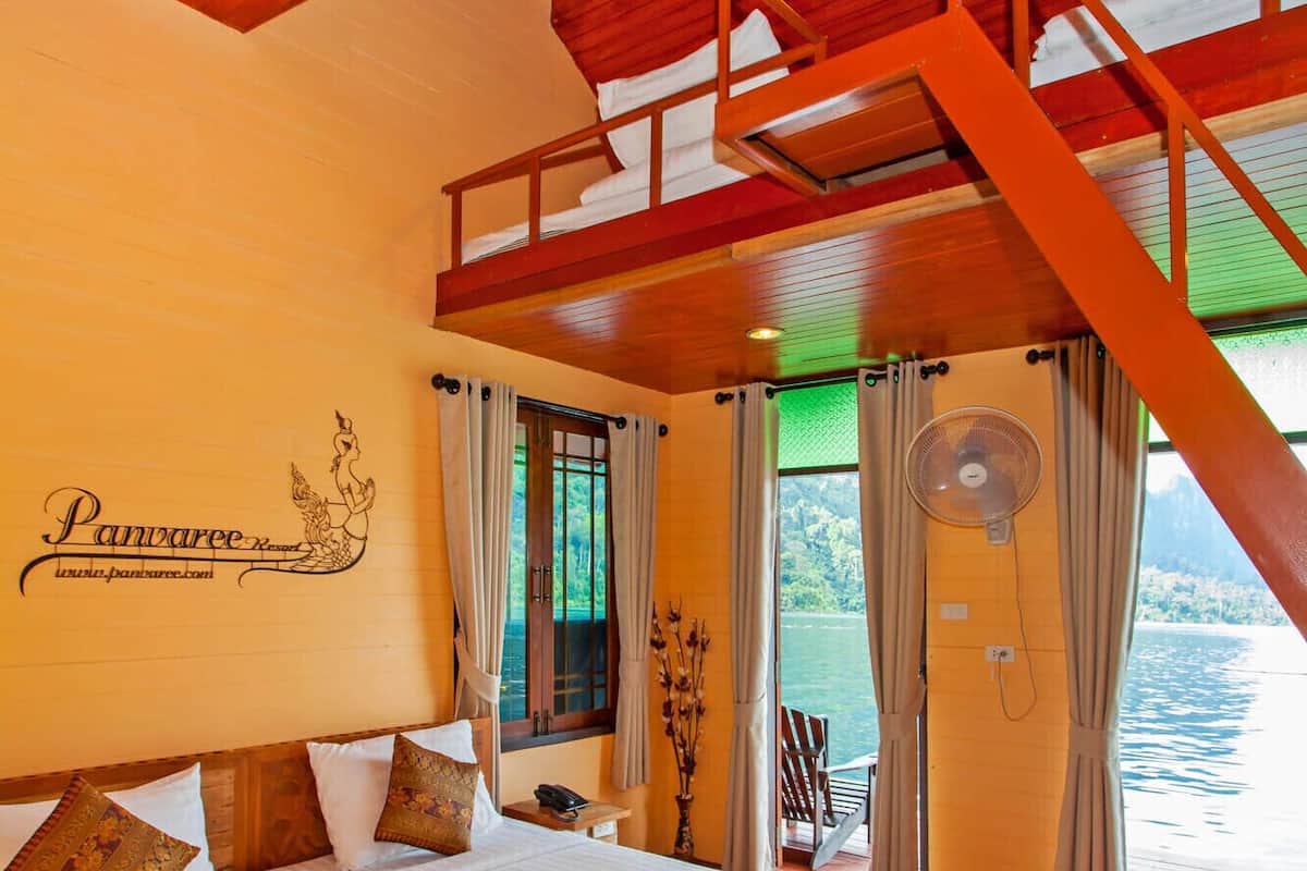A bedroom with a wooden ceiling, a bed with white linens, a nightstand, a fan, and a view of the water outside.