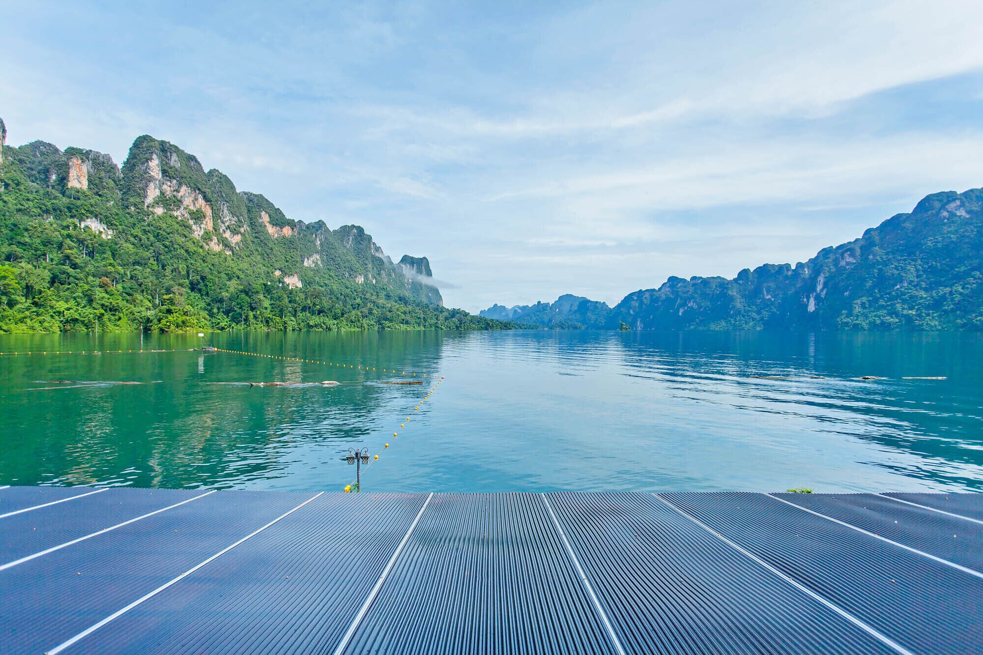Convés de barco com painéis solares, água calma e falésias arborizadas.