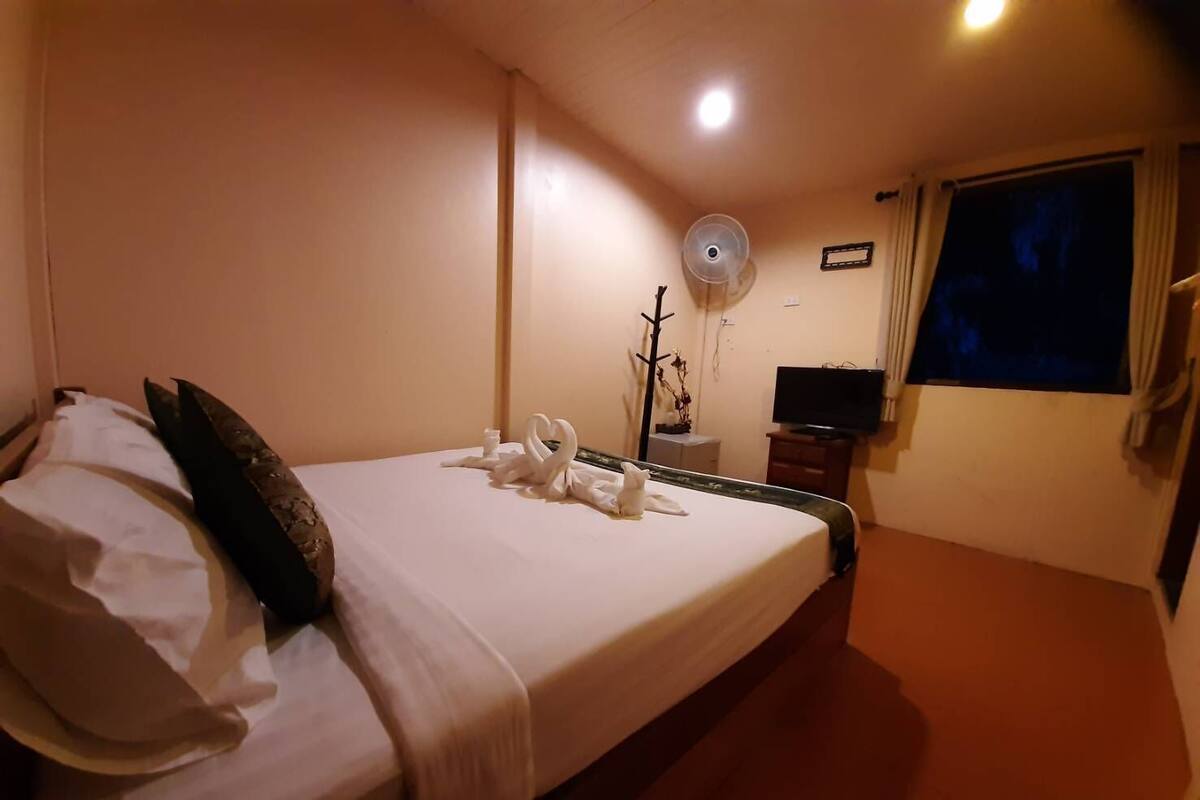A hotel room with a neatly made bed, a flat-screen TV, a standing fan, and a window with curtains.