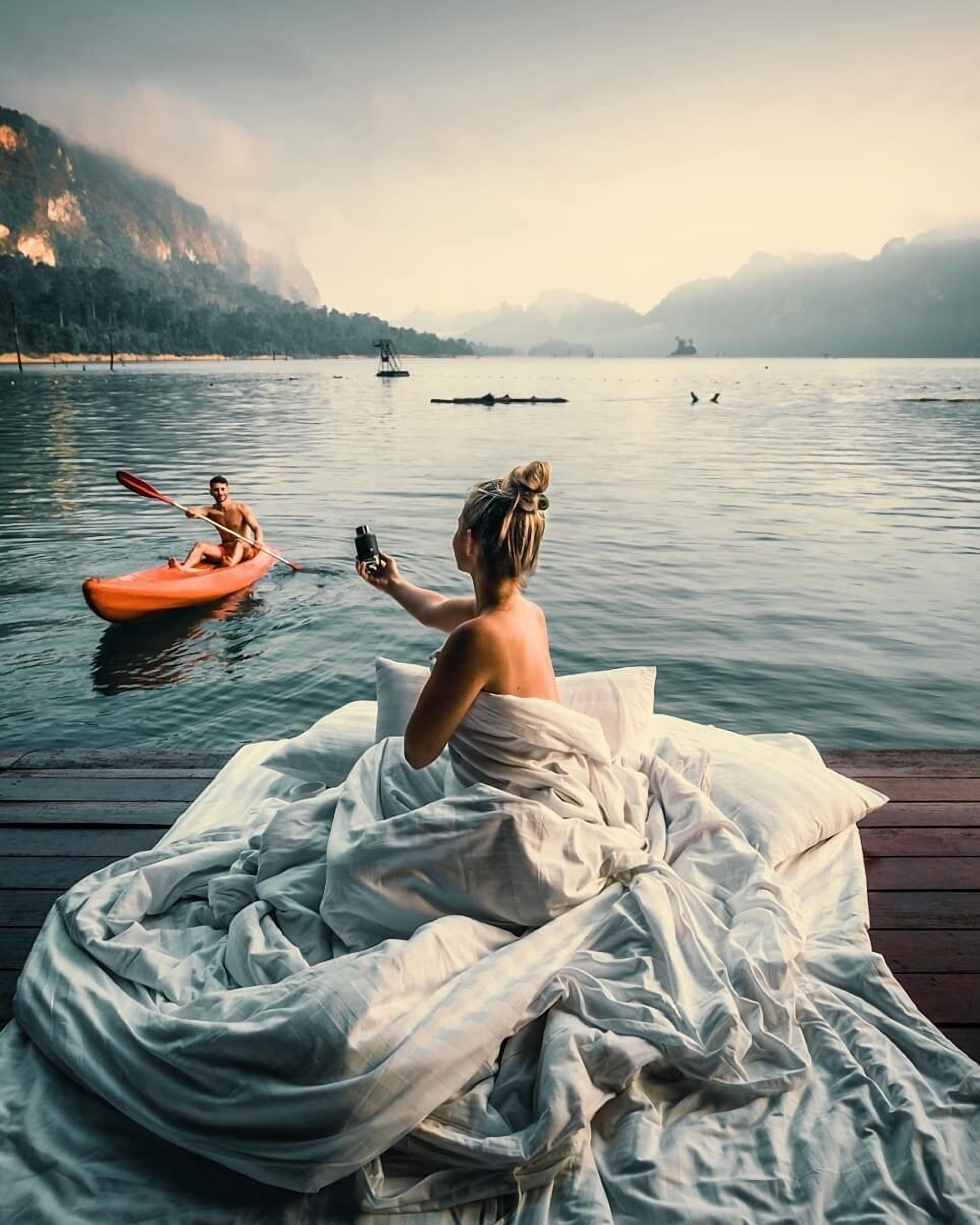 A person in a white dress sitting on a wooden dock by a calm lake with mountains in the background.