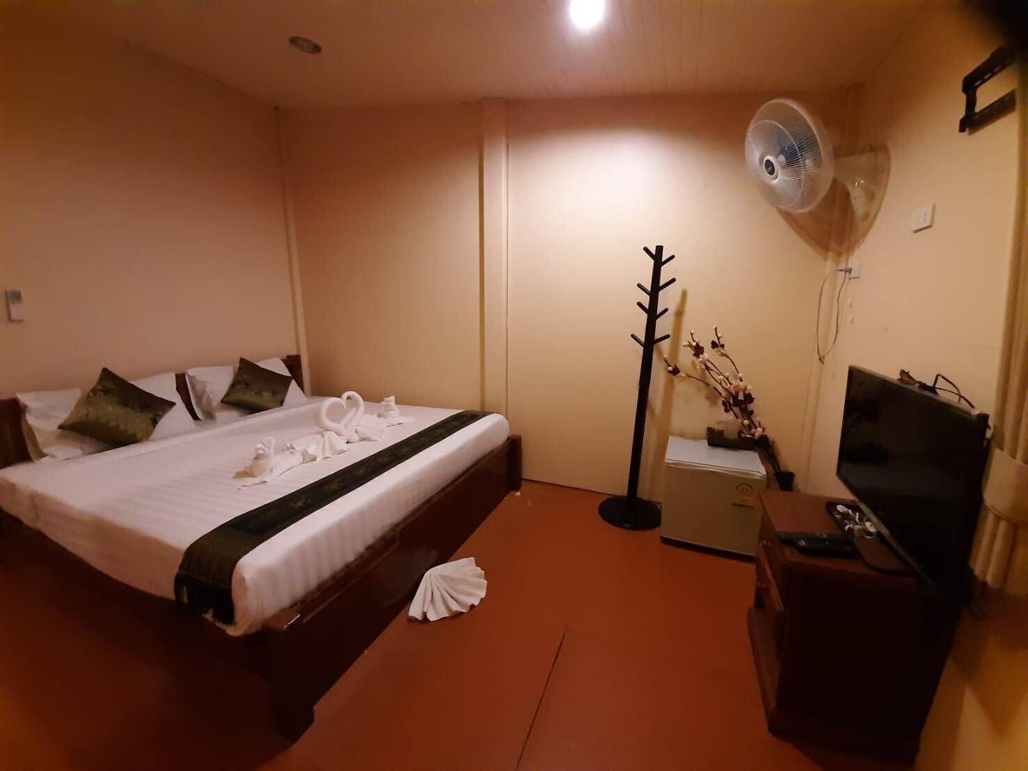 A hotel room with a bed, a desk with a television, a fan, and a decorative plant.