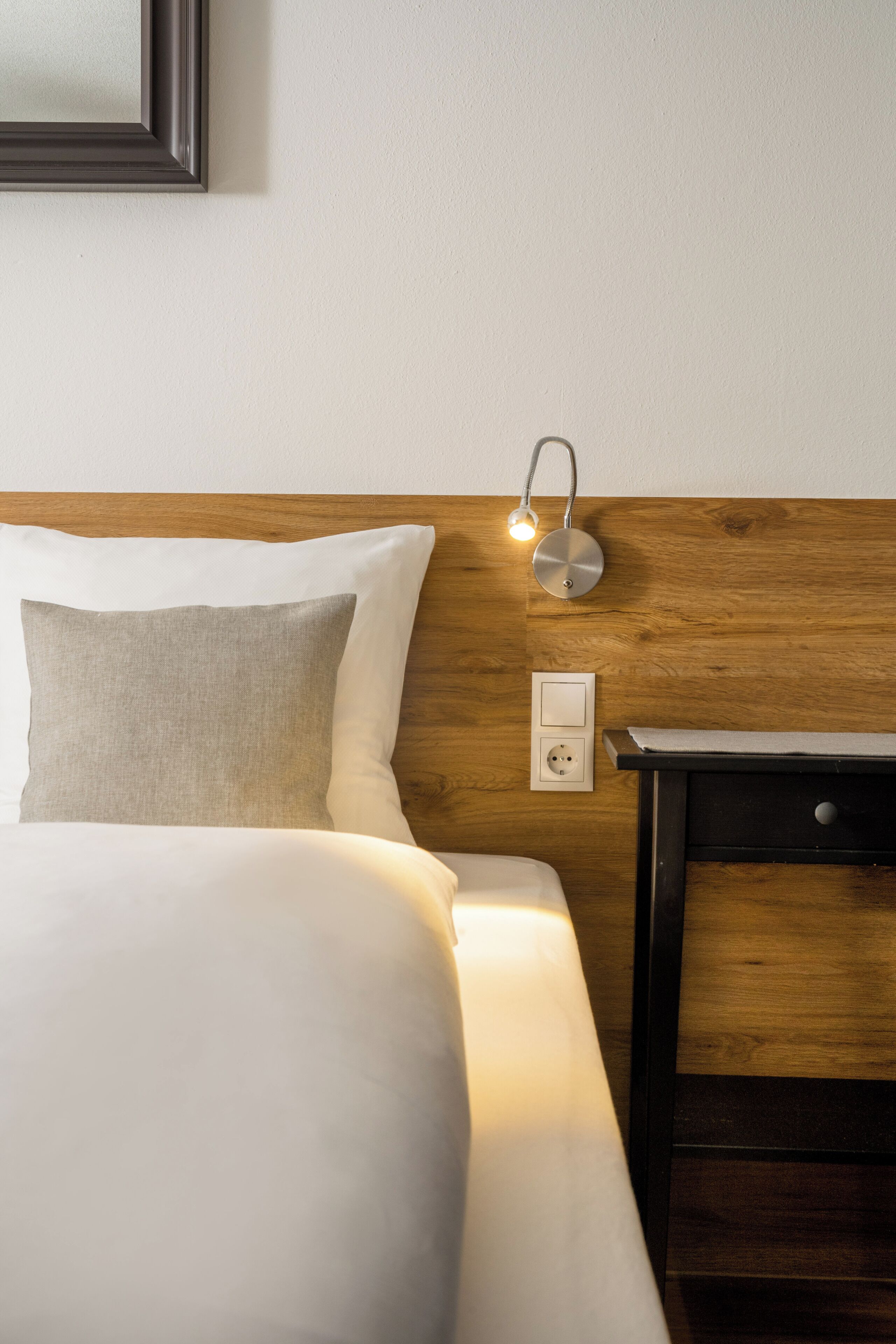 A neatly made bed with white linens, a wooden headboard, a bedside table with a lamp, and a wall socket.