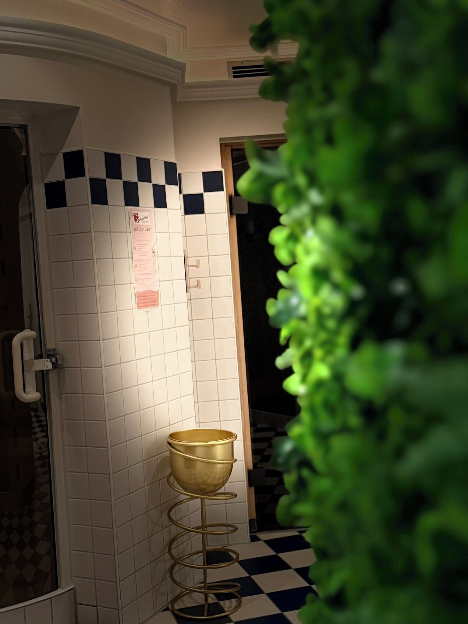 Een badkamer met witte betegelde muren, een zwart-wit betegelde vloer, een gouden emmer op een spiraalvormige standaard en een deur met een knop.