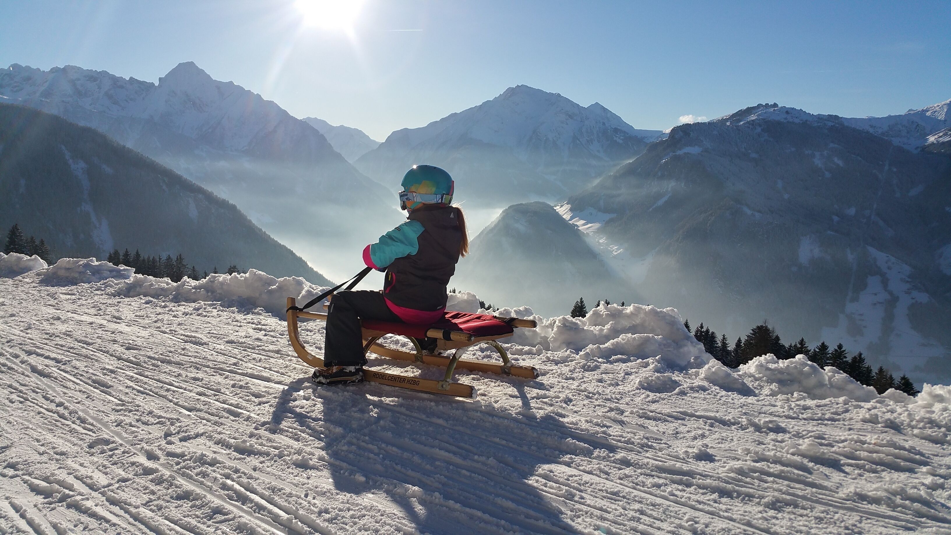 Une personne sur une luge, sur une pente enneigée d’une montagne, avec un ciel dégagé et des montagnes au loin.