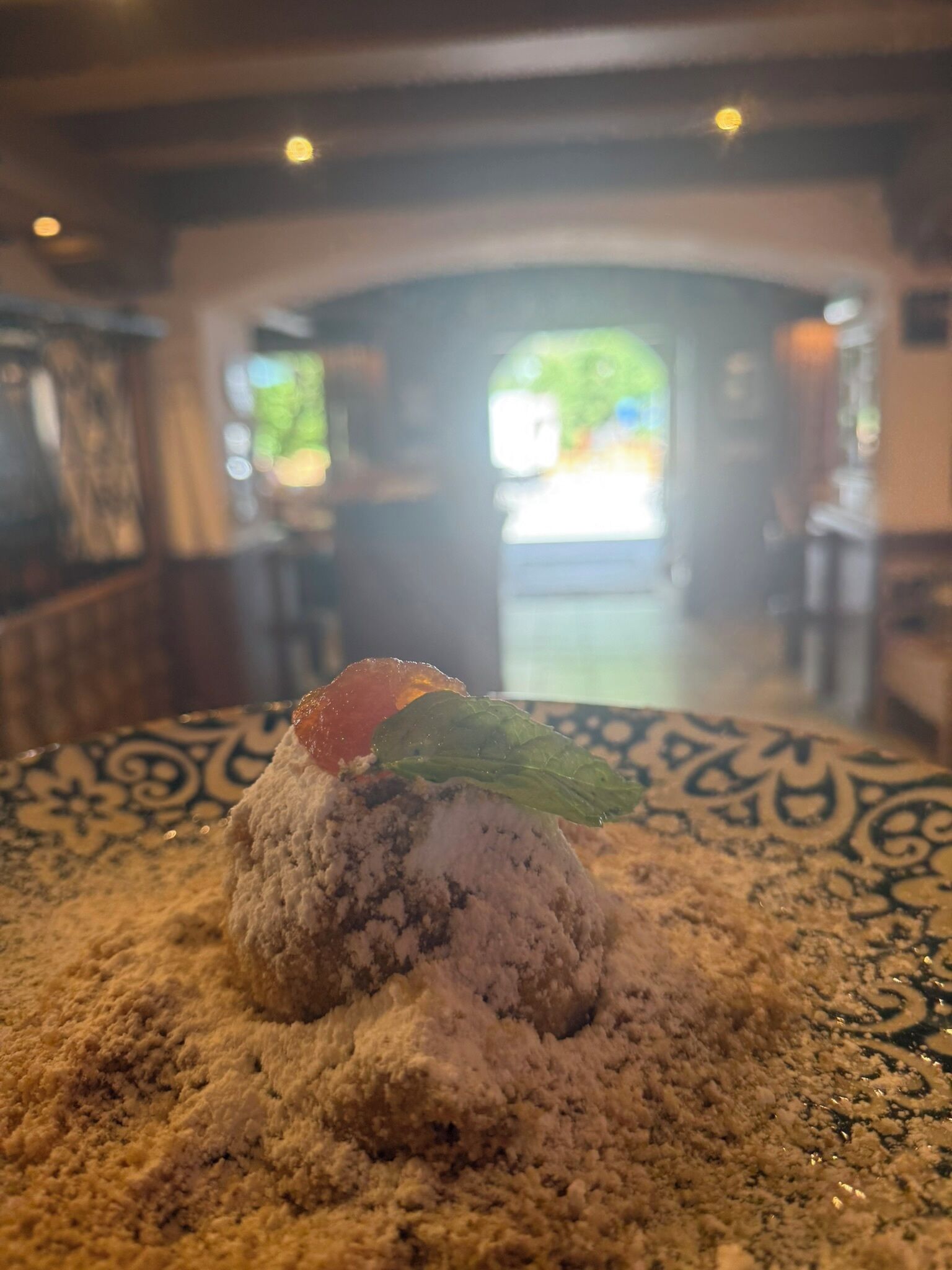 Un postre en un plato con un diseño decorativo, situado en un restaurante con vistas a un espacio exterior verde de fondo.