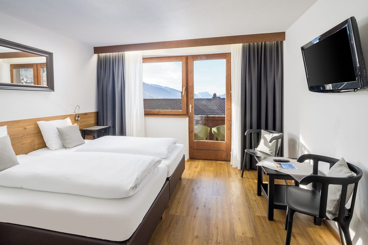 A hotel room with a bed, a desk with a chair, a TV, and a view of mountains through a window.